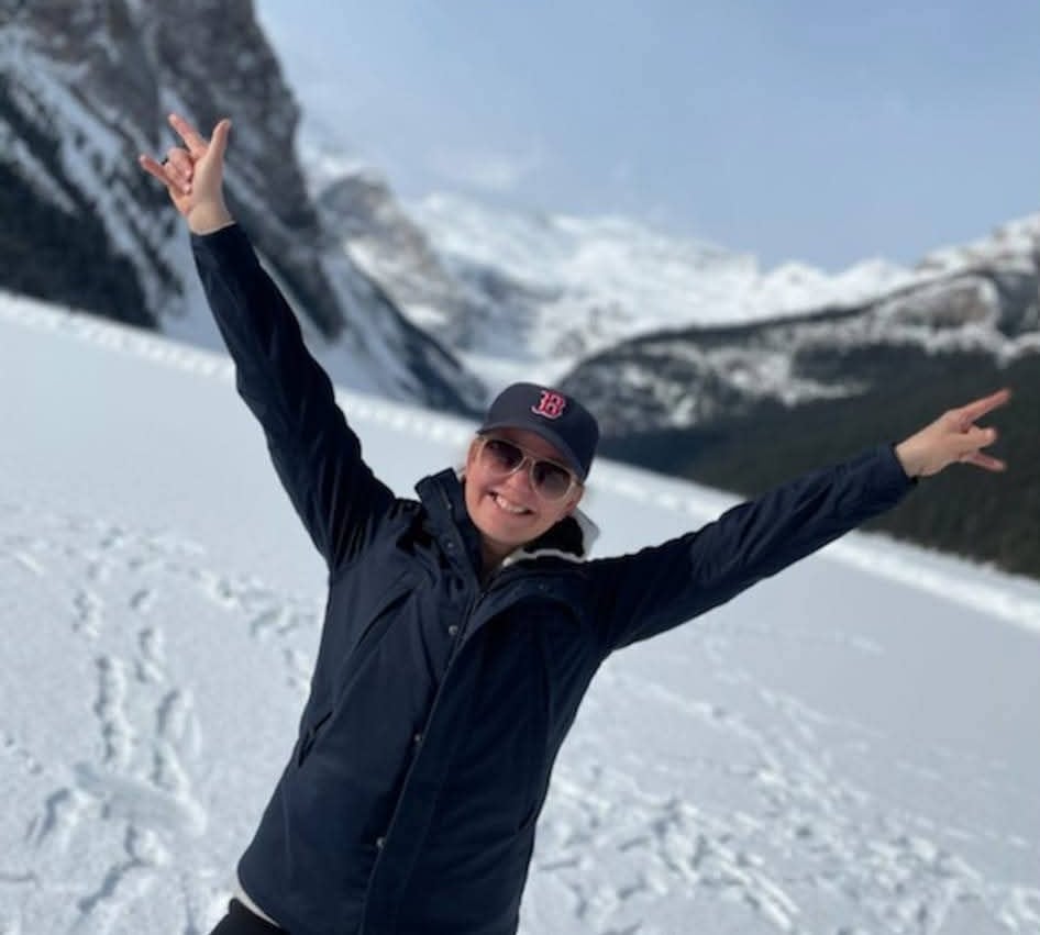 A smiling woman happily raises her arms while standing in front of a frozen lake and snowy mountains.
