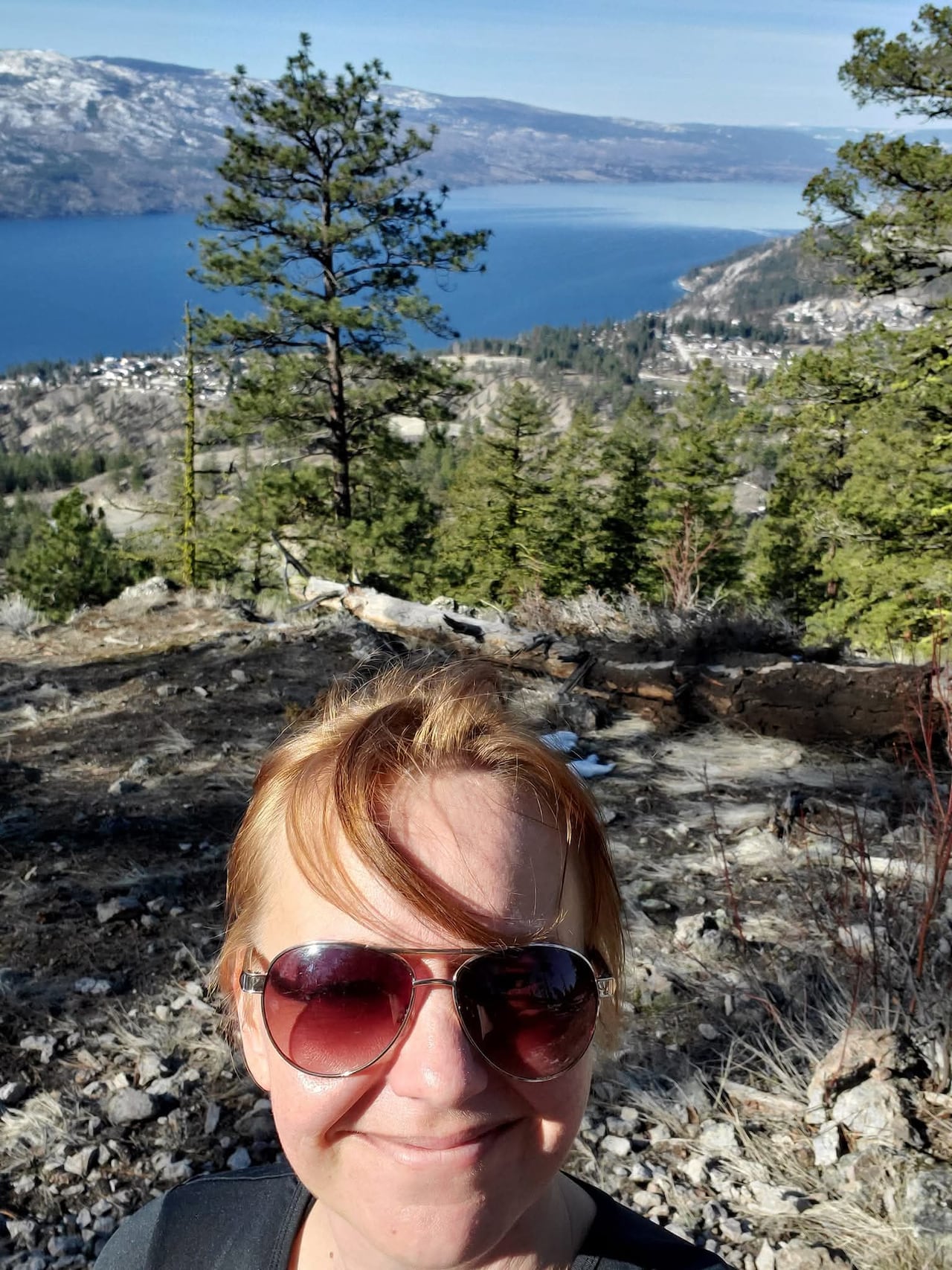 A smiling woman stands on a mountain ridge overlooking a lake. 