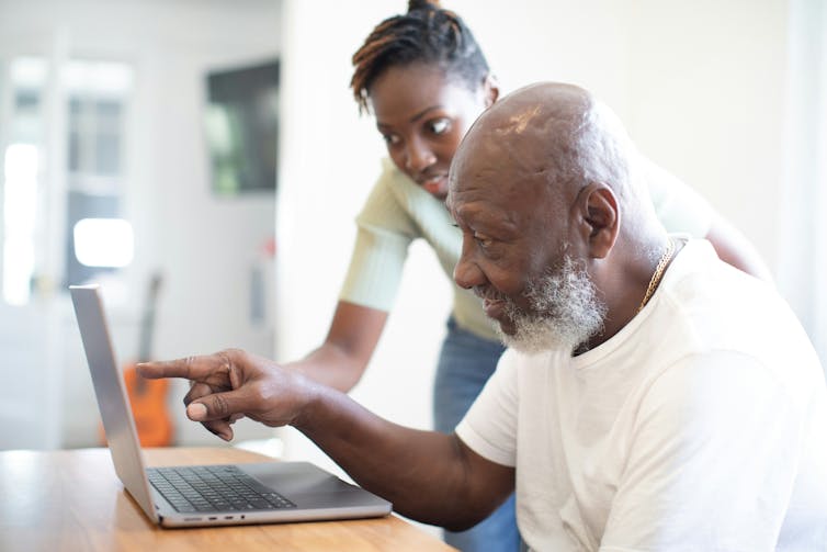 A man with a gray beard is pointing at a laptop screen while a young woman is looking over his shoulder.