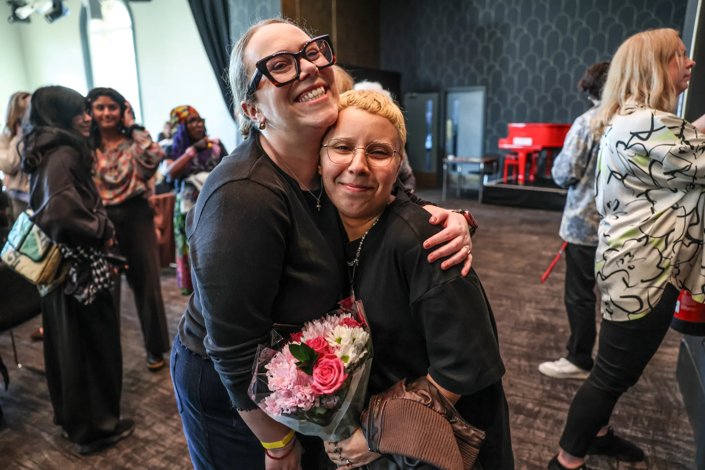 Carly Smallman hugs a woman with short red hair holding a bouquet of flowers, after a stand-up comedy performance.