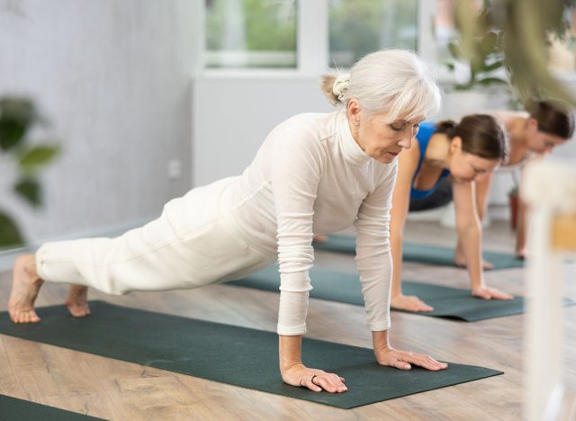 Concentrated elderly woman holds plank pose to strengthen body muscles during group yoga training in studio. Core exercises for older adults. The concept of an active lifestyle.