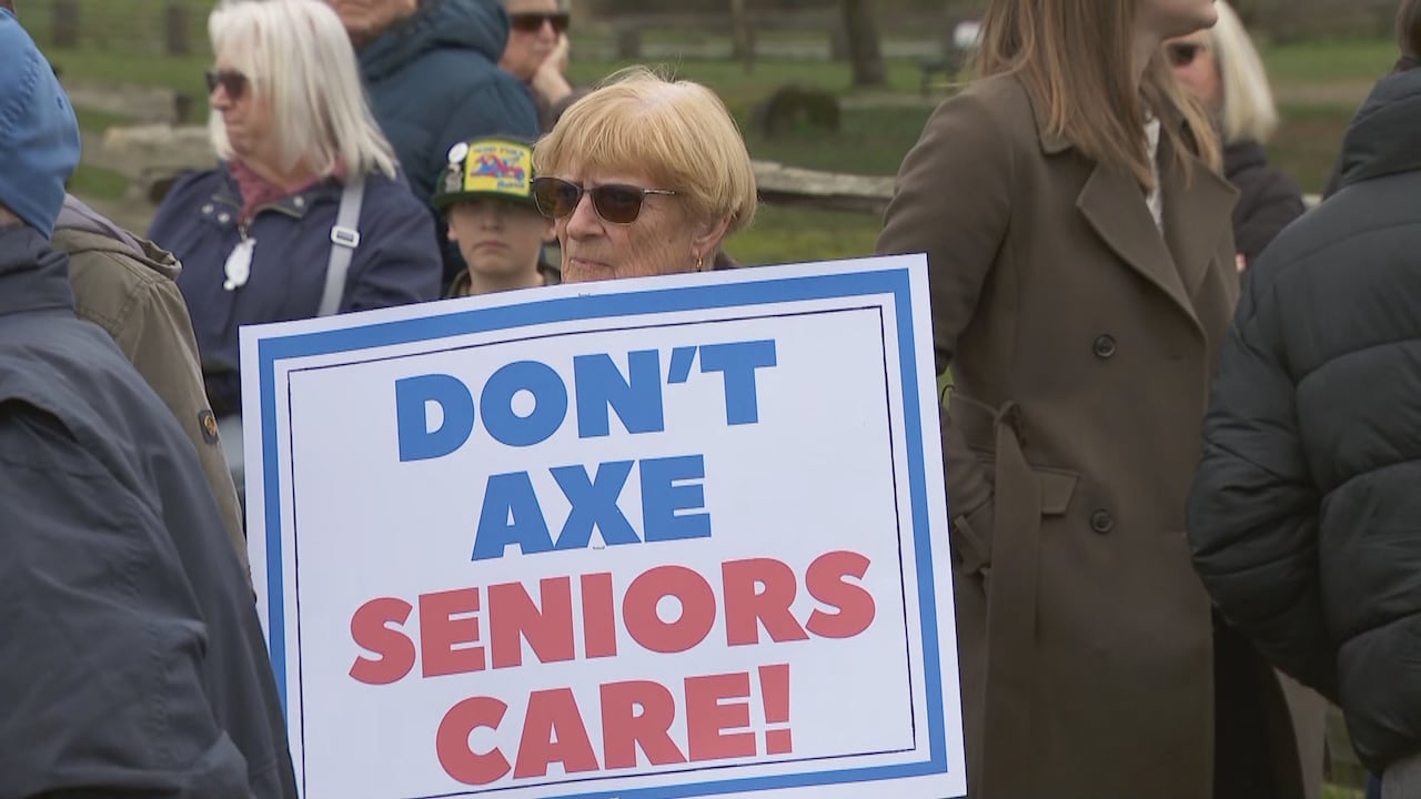An elderly woman wearing sunglasses holds a sign while standing among a group of people outdoors.