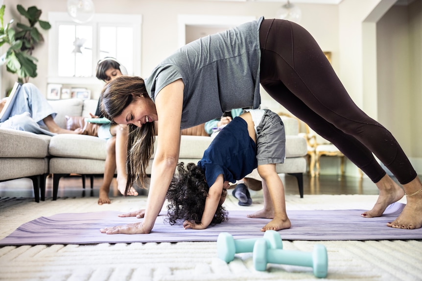 The mother is exercising at home while being distracted by the little boy.