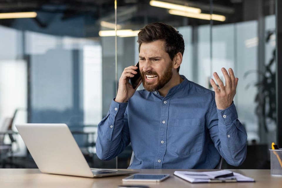 Angry and angry young businessman sitting at desk in office and talking on phone while raising hands