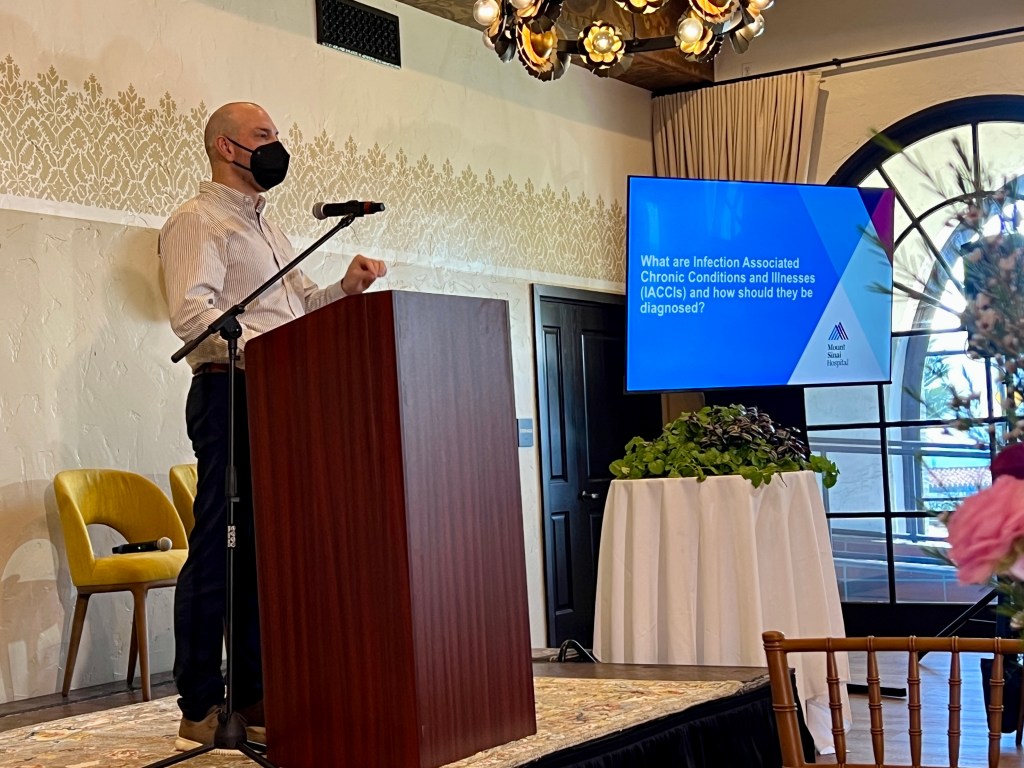 David Putrino speaks backstage at the lectern while giving a keynote address about chronic COVID and IACCs. He is a white man with a shaved head and a white button-up shirt and black breathing. Next to him on the screen is a slide from his presentation that reads, "What are the chronic conditions and diseases of infection and how should they be diagnosed?"