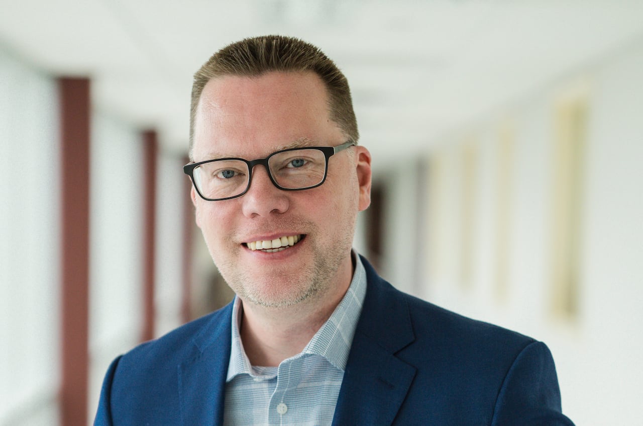 Professional portrait of a man with short brown hair and glasses, wearing a blue jacket and checked shirt.