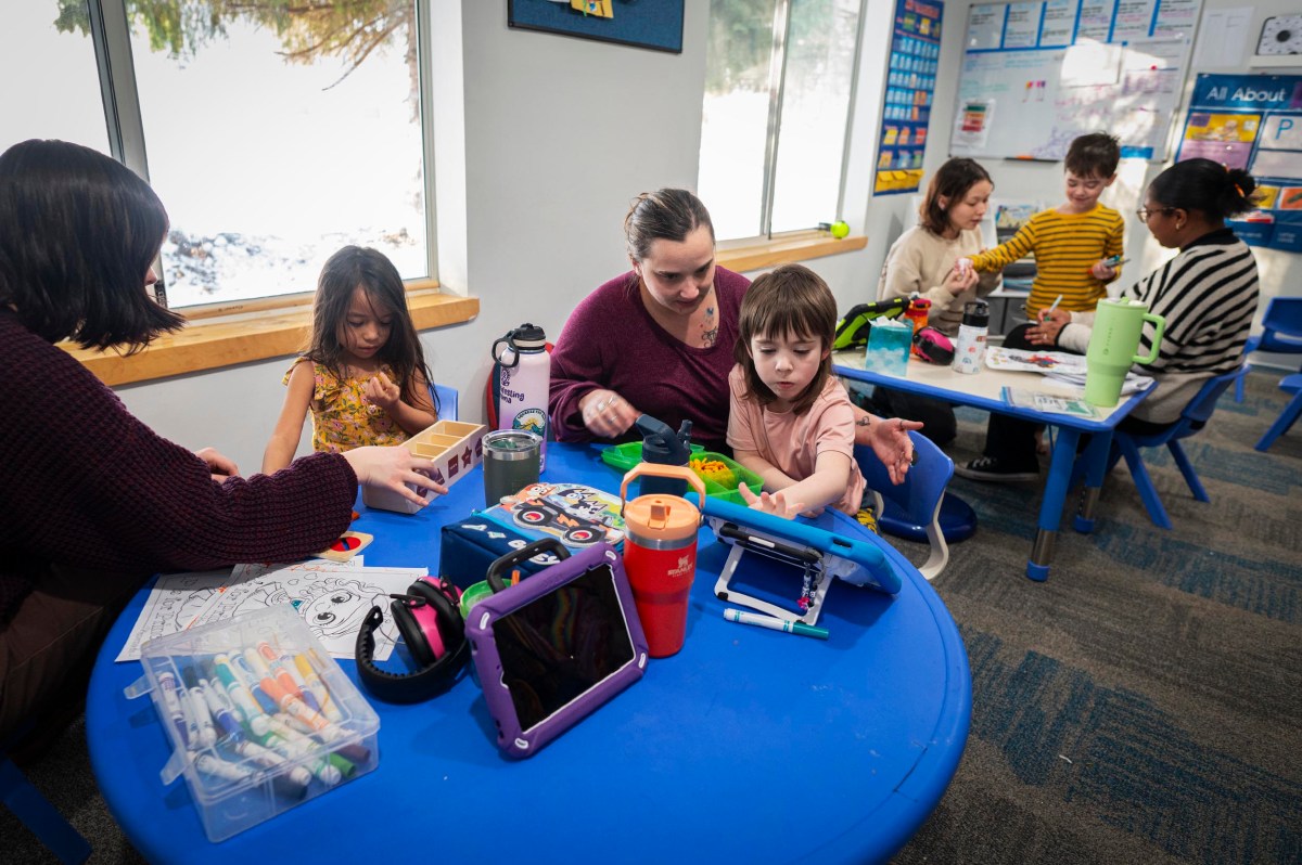 Children and adults sit at blue tables in the classroom, engaged in activities with tables, coloring supplies, and educational materials.