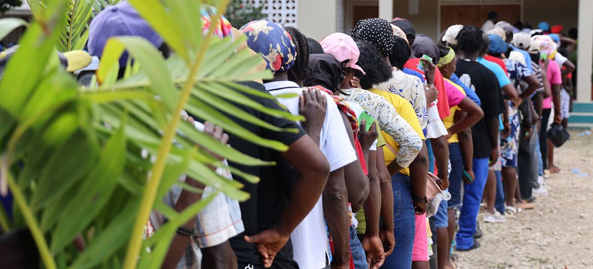 A long line of people wait outside for cash distributions as part of the World Food Program (WFP) 'Cash for Work' program in Haiti, with green tropical plants in front.
