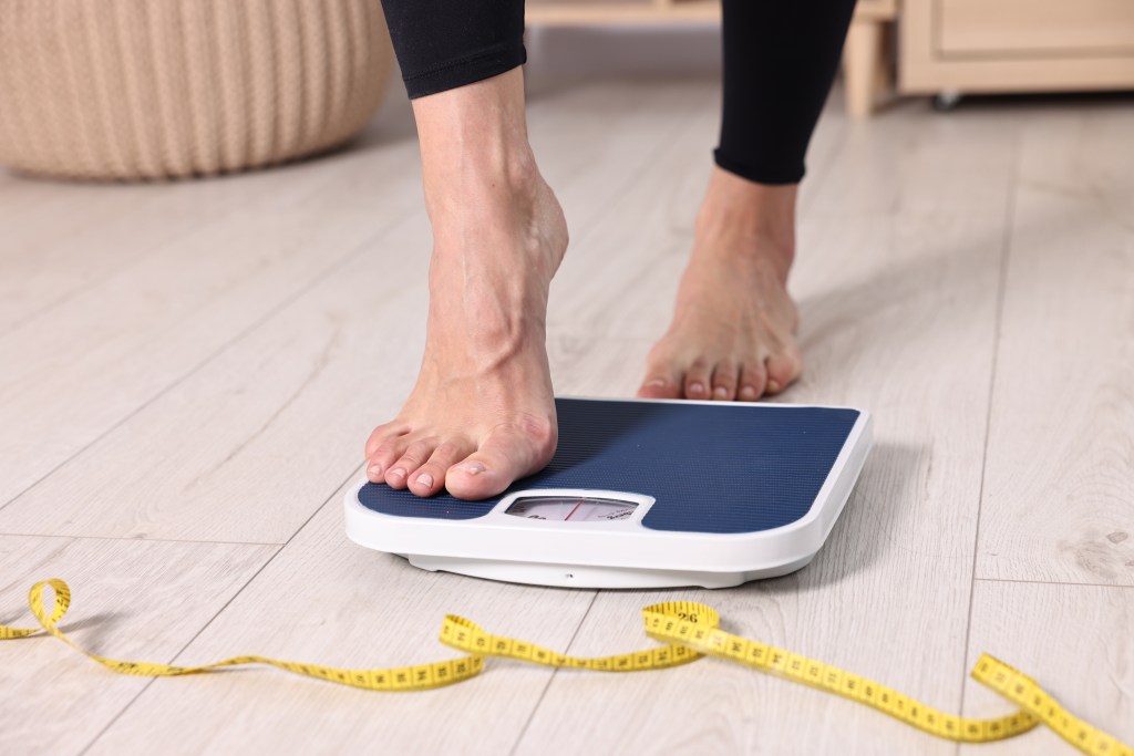 A woman steps on a bathroom scale, with a yellow measuring tape on the floor beside her.