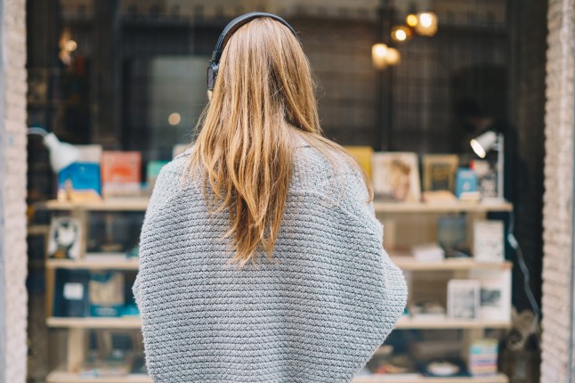 Young Caucasian woman in back with headphones and pullover looking at bookstore window.