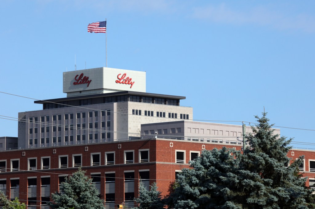 The exterior of the Eli Lilly & Co. headquarters building in Indianapolis, Indiana, with the Lilly logo on the roof and an American flag flying overhead.